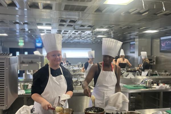 Image depicts two young people wearing chef hats and white aprons cooking in the kitchen.