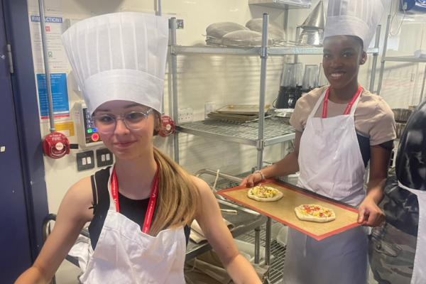 Image depicts two young people holding baking trays of pizza.