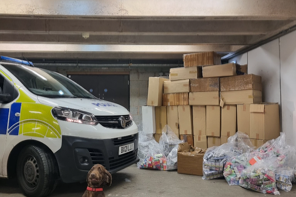 Image depicts a sniffer dog in front of seized goods and a police van.