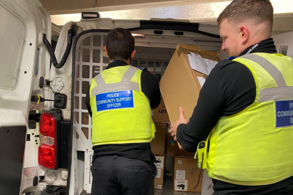 Image depicts two police community support officers placing seized boxes in the back of a police van.