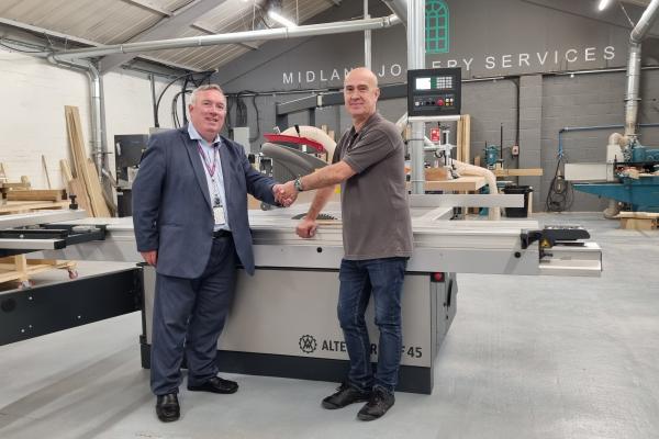 Two men shake hands, in front of a large piece of machinery, and smile at the camera