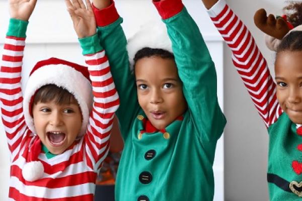 three young children in elf costumes and festive Santa hats with their arms in the air.