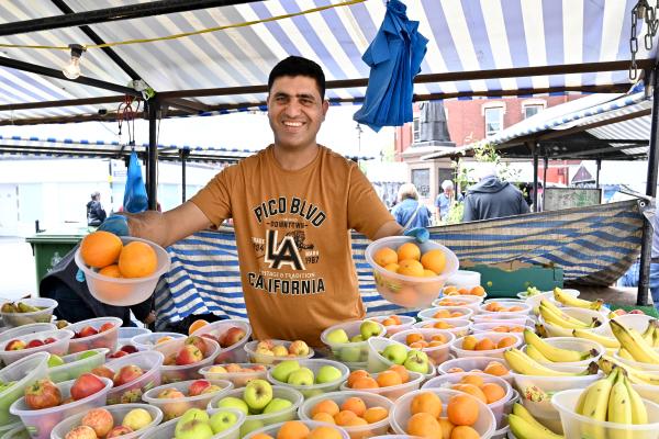 A smiling trader at their stall on Walsall Market holding bowls of oranges and surrounded by different fruit in bowls