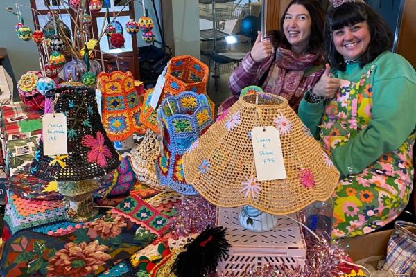 Two stallholders at a craft fair, with hand crafted items laid out on the table in front of them.