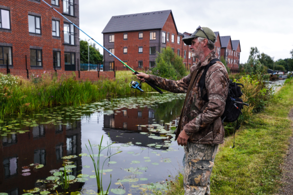 Image depicts a man fishing by the canal with houses in the background.