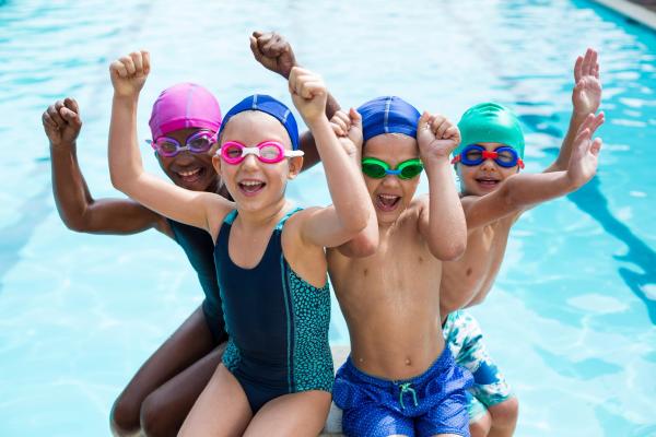 group of four children in swimming costumes with hands in the air cheering with a swimming pool background