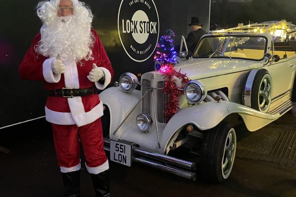 Image depicts Santa Claus in front of a decorated vintage car for a Christmas festival.