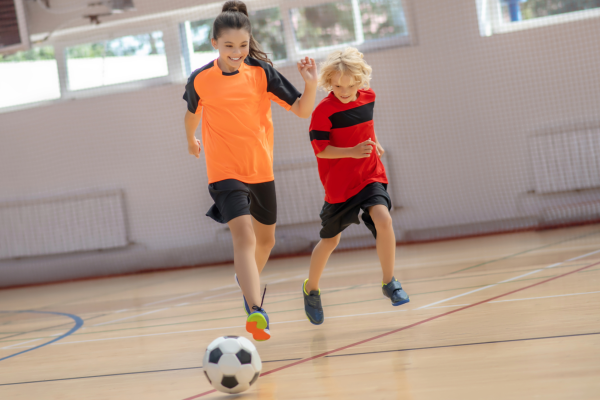 boy and girl kicking a football in an indoor sports hall smiling