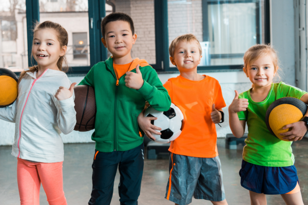 young children holding football, basketballs in an indoor sports hall with windows in the background 