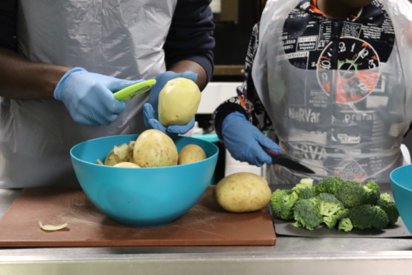 Image depicts two people peeling potatoes and chopping broccoli. 