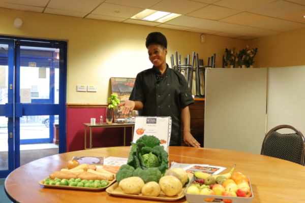 Image depicts a chef in chef blacks presenting a variety of fruit and vegetables.