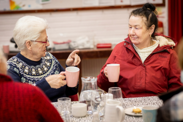 Image depicts two women of older age enjoying a warm drink in a community setting.