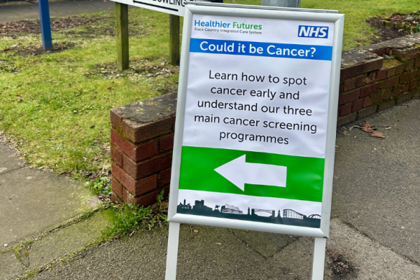 Image depicts a sign outside a community centre that reads 'Could it be Cancer? Learn how to spot cancer early and understand our three main cancer screening programmes'