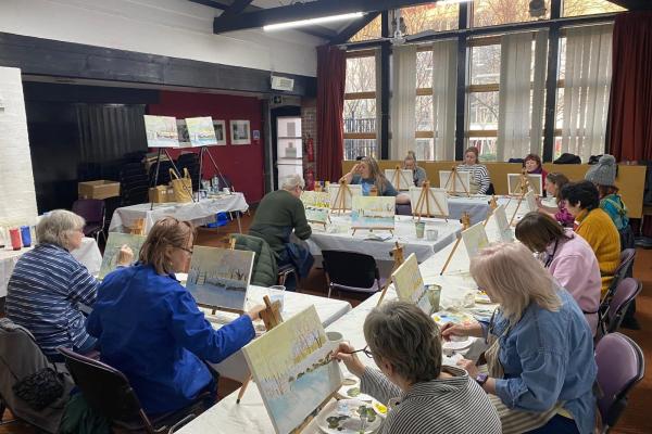 Group of people in a large room with painting materials, easels, canvas, paints etc