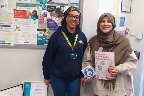 Image depicts two women holding a sticker and poster for breastfeeding friendly scheme.