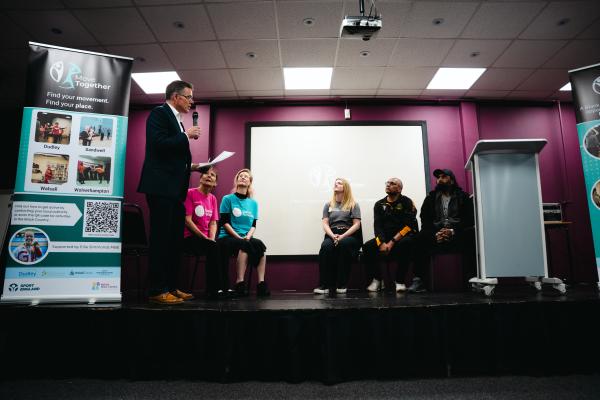 Image depicts a panel discussion of five people and a presenter at the Move Together campaign launch.