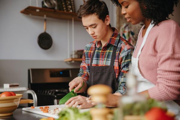 Image depicts a woman helping a young person with basic knife skills in the kitchen.