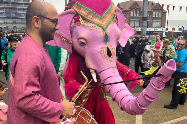 A man playing a bhangra drum with a human sized pink elephant puppet