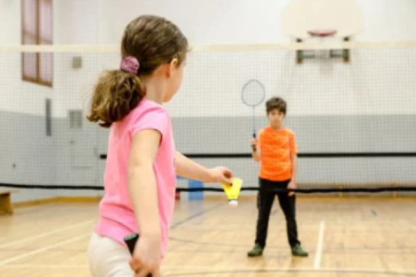 young girl in a pink top and blurred out boy in an orange top playing badminton in a sport shall 